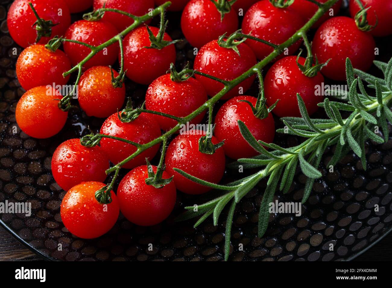 Pomodori ciliegini rossi maturi umidi sulla vite e sprig di rosmarino fresco su un piatto nero. Macro. Gocce d'acqua su un piccolo pomodoro rotondo. Freschezza, saporita Foto Stock