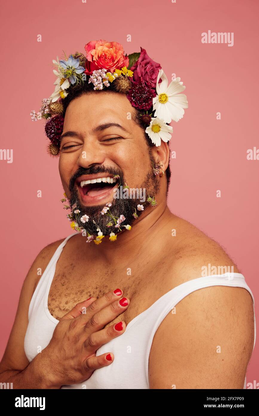 Queer sorridente che indossa il trucco e il canotta. Allegra drag queen indossando fiori in testa e barba. Foto Stock