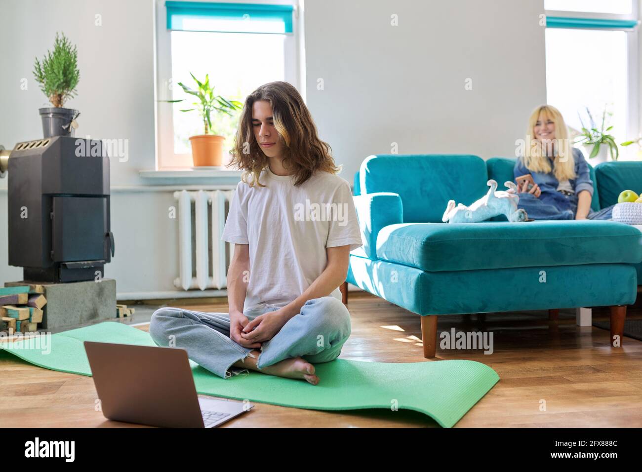 Ragazzo teenager seduto in posizione di loto su tappetino yoga a. casa a pavimento con computer portatile Foto Stock