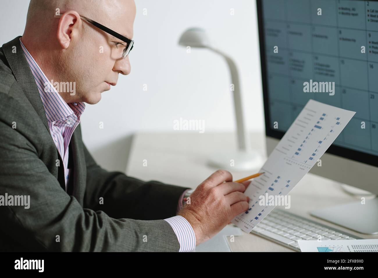 Concentrato uomo d'affari maturo in occhiali seduto alla scrivania con computer e visualizzazione di statistiche su carta Foto Stock