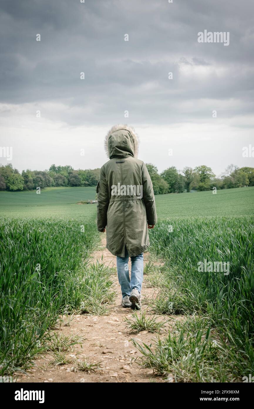 Vista posteriore di una donna che indossa un cappotto Parka che cammina lungo una pista in un campo di grano con un cielo tempestoso. Foto Stock