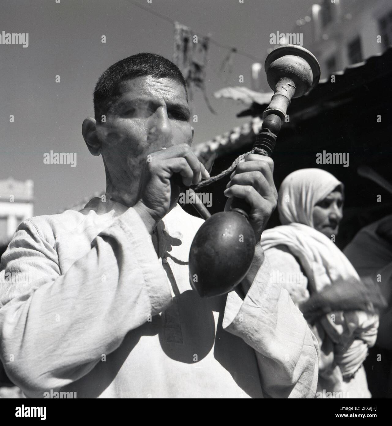 Anni '50, storico, uomo che fuma o che crea fumo da un lungo, strano tubo di legno, India. Con un'estremità in gomma e un piano circolare in legno, è un oggetto inusal, forse usato in rituali e cerimonie religiose. In diverse culture, un tubo cerimoniale creerebbe fumo per portare preghiere all'attenzione del creatore (Dio) e di altri spiriti. Qui l'uomo sta forse conducendo un entourage e/o una processione di nozze attraverso la città e creando il fumo per chiedere la coppia sposata per essere benedetto. Foto Stock