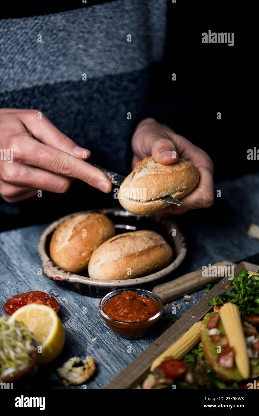 un giovane caucasico taglia un panino per preparare un panino o degli spuntini vegani, seduti ad un tavolo di legno grigio, accanto ad un vassoio con un po' di appe vegano Foto Stock