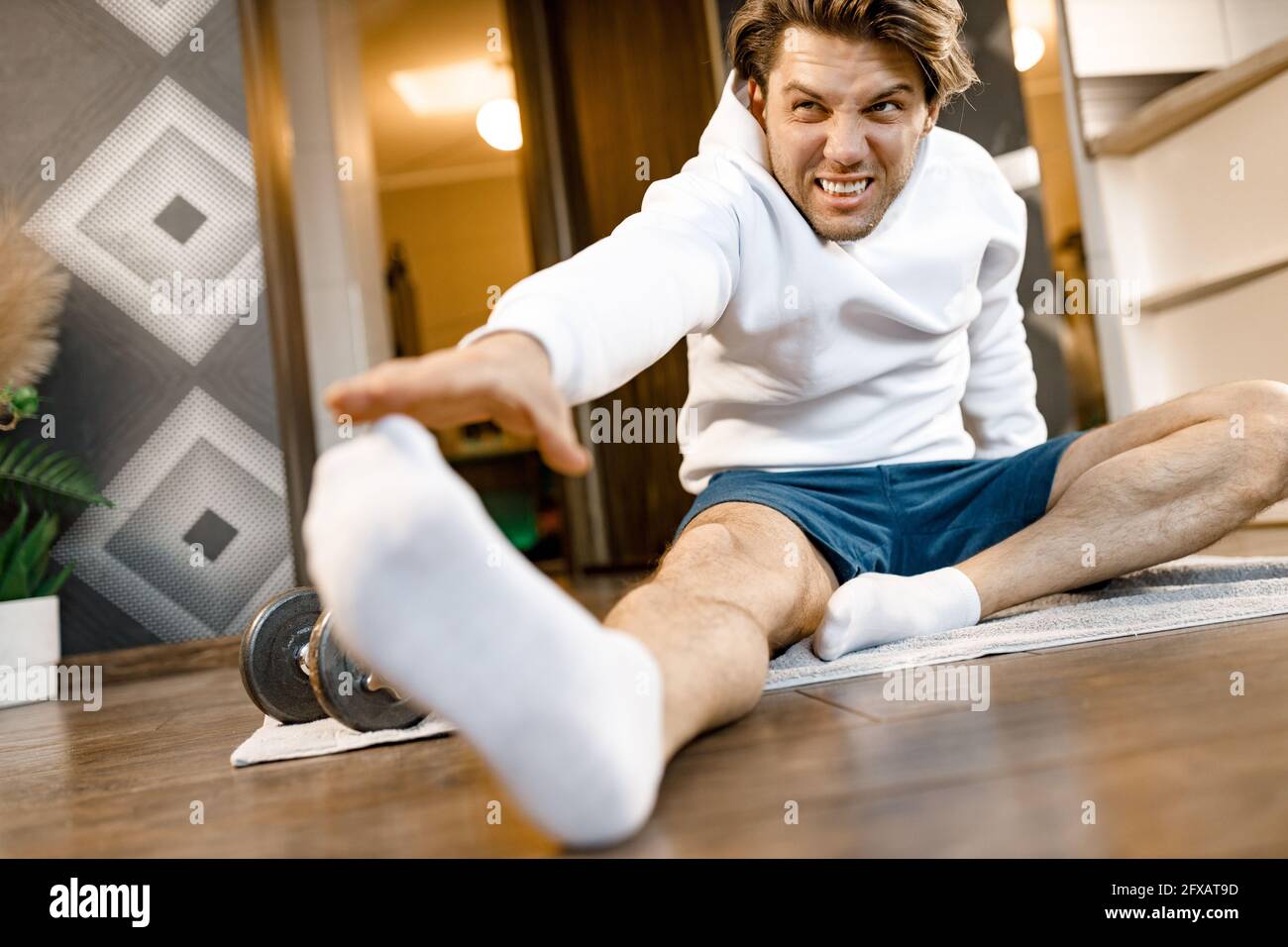 Bel maschio sportivo seduto sul pavimento, facendo stretching dopo l'allenamento. L'uomo stanco si allenava a casa mentre era in quarantena, mantenendosi in buono Foto Stock