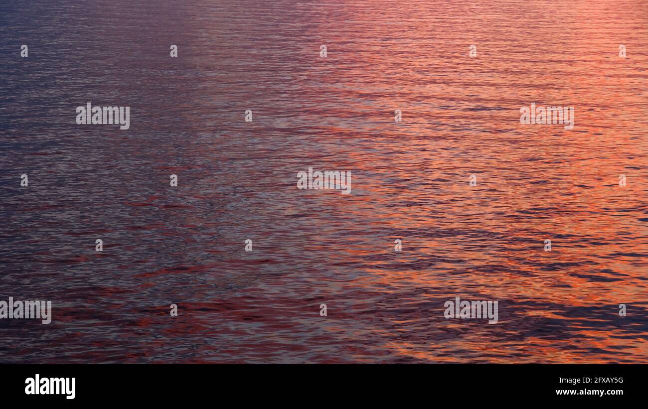 Una superficie d'acqua di tre colori arancio, rosa e blu scuro con increspature e riflessi soleggiati in mezzo al mare. Tramonto o alba riflesso d'acqua Foto Stock