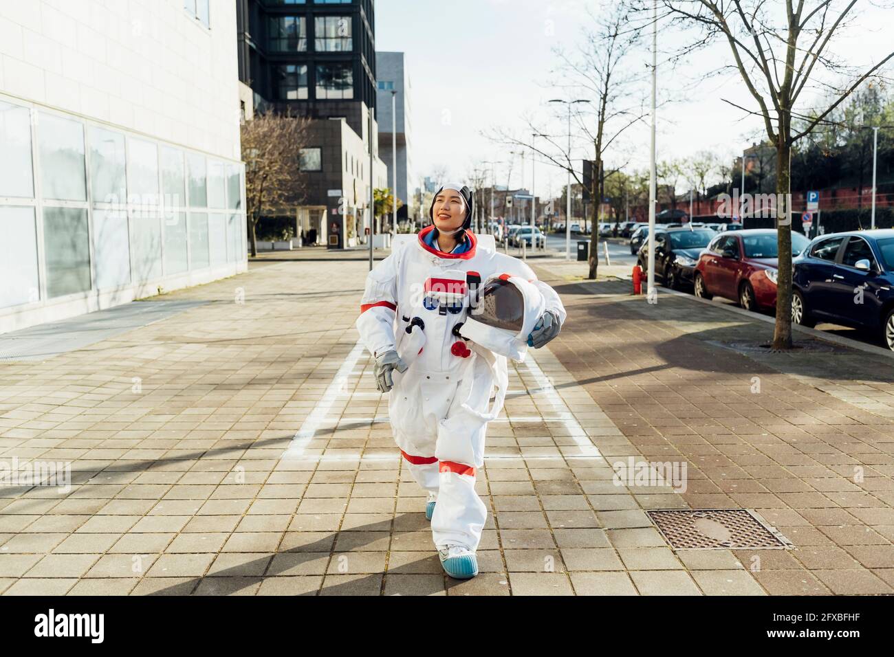 Sorridente astronauta femminile che tiene il casco spaziale mentre cammina sul sentiero in città Foto Stock