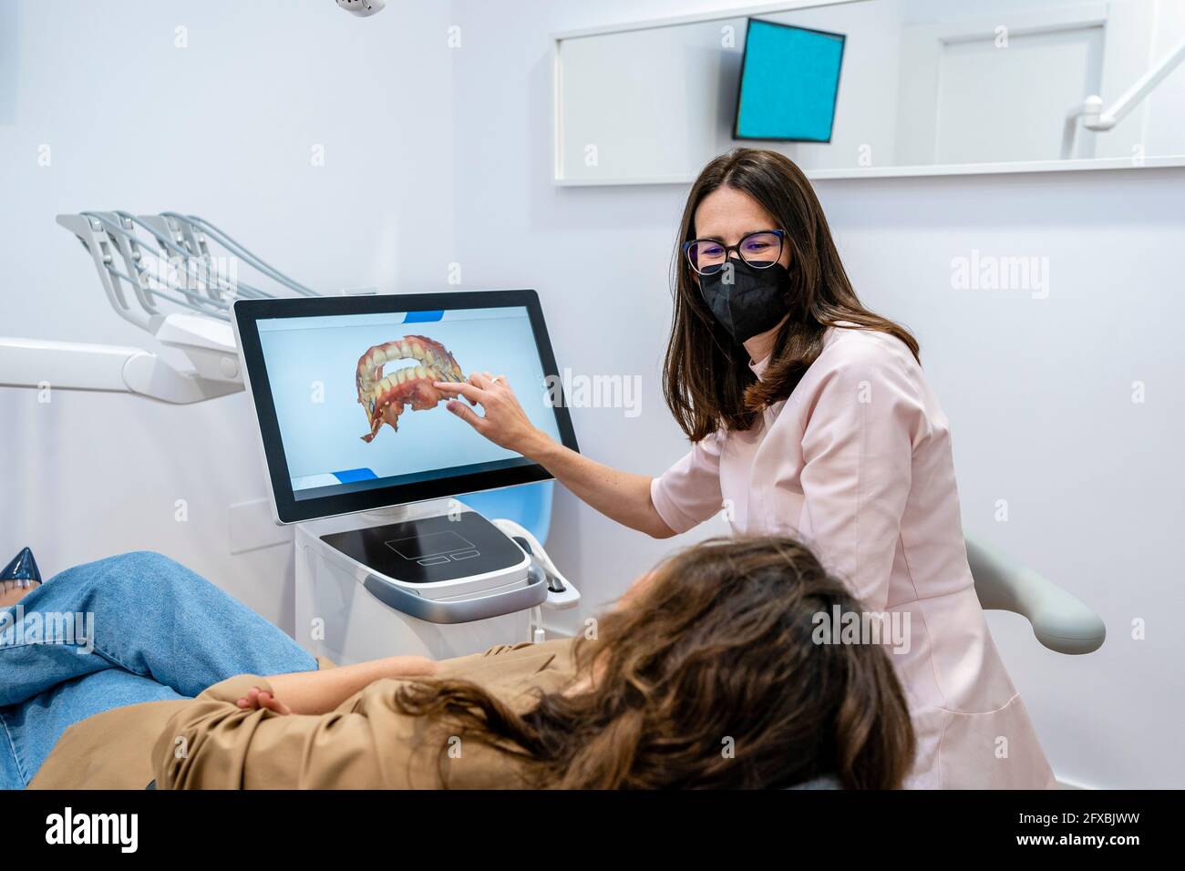 Dentista femminile che indossa una maschera protettiva che spiega sullo schermo del computer al paziente in clinica medica Foto Stock