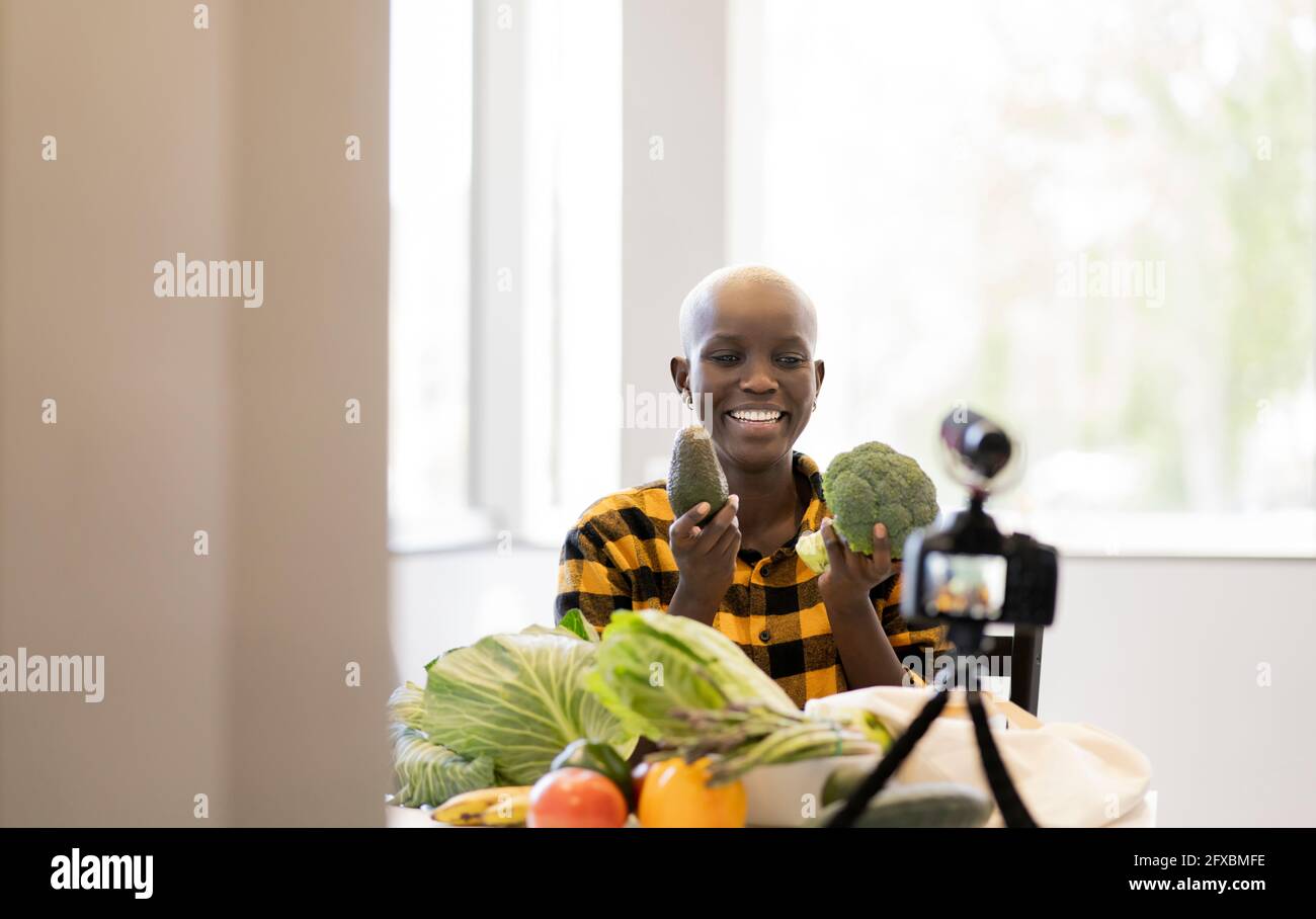 Sorridente influencer femminile che mostra vegatbles mentre vlogging usando la videocamera a casa Foto Stock
