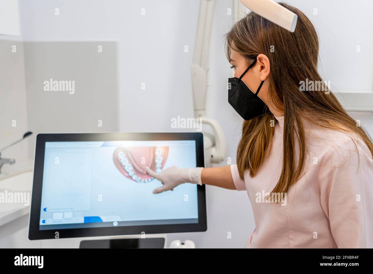 Dentista femminile che indossa una maschera protettiva rivolta verso lo schermo del computer in clinica Foto Stock