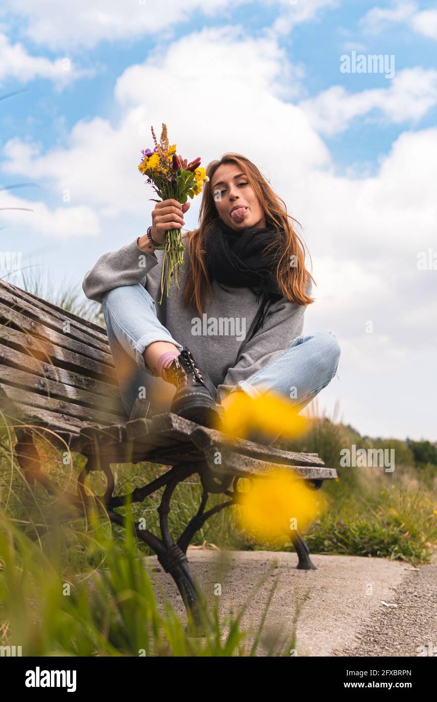 Giovane donna che si stacca la lingua mentre si siede con mazzo di fiori sulla panchina Foto Stock