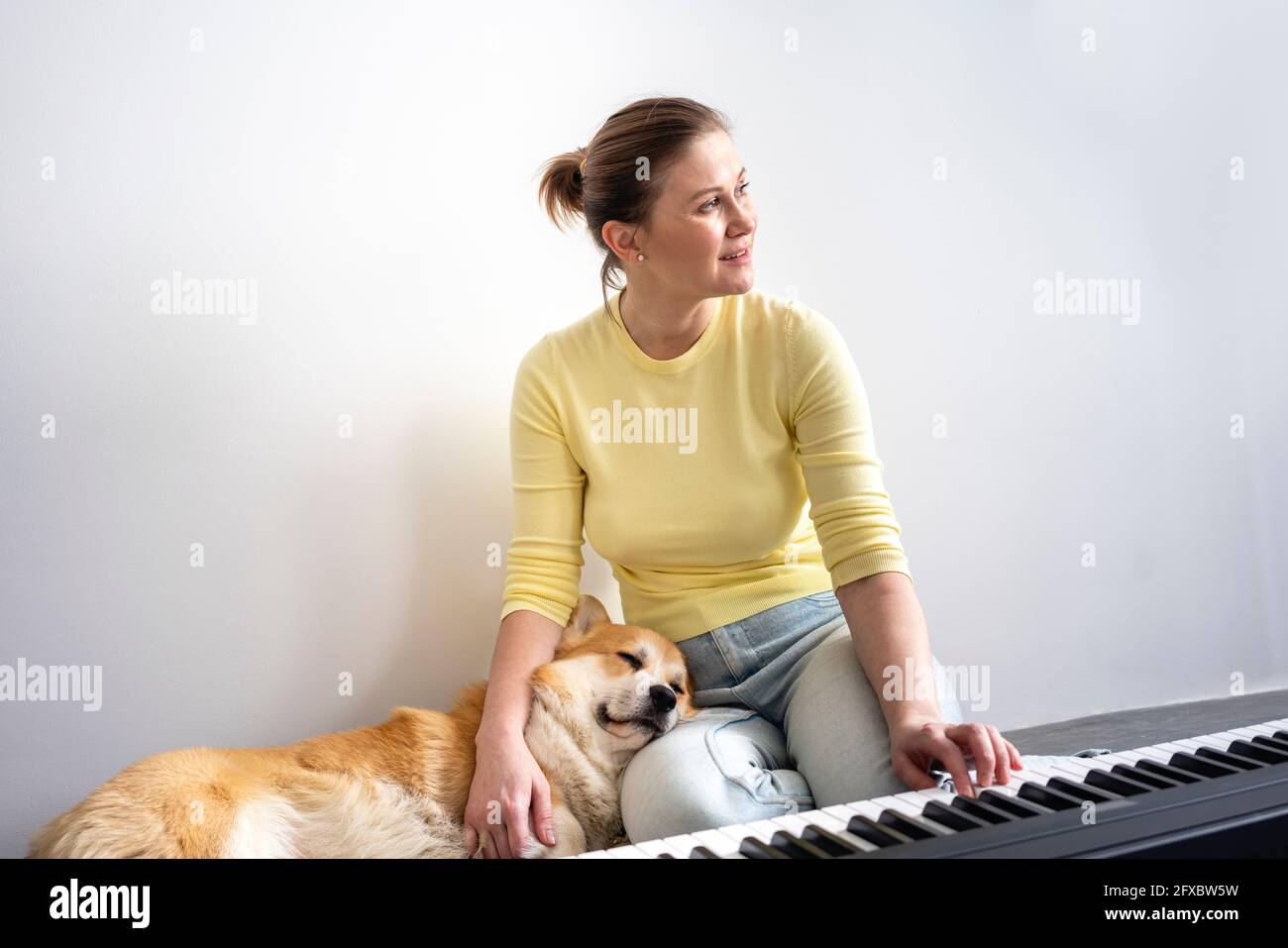 Donna sorridente che guarda via mentre il cane dormiva sul grembo davanti al pianoforte a casa Foto Stock