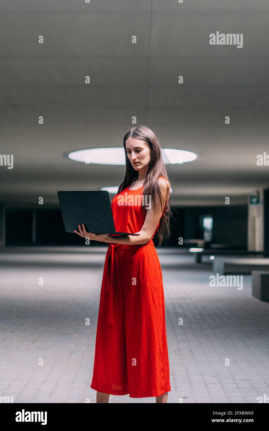 Donna d'affari in abito rosso, in piedi lavorando con il computer portatile in mano all'interno. Foto Stock