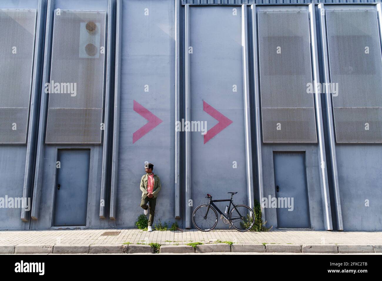 Uomo con la bicicletta sul sentiero che guarda lontano mentre si appoggia sulla parete della fabbrica Foto Stock