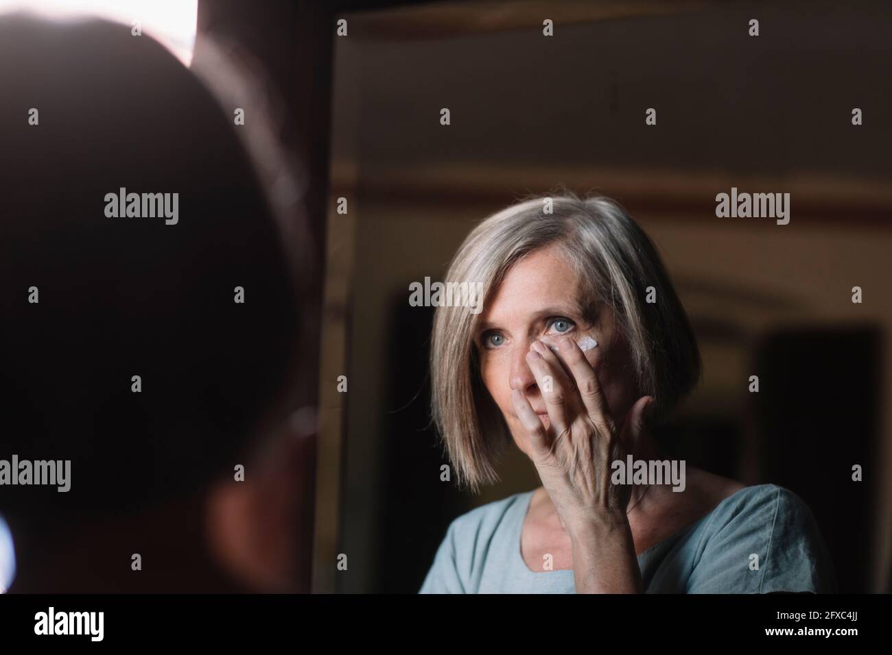 Donna dai capelli corti che guarda nello specchio mentre applica la crema del viso a casa Foto Stock