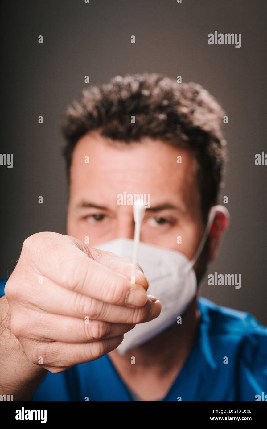 Operatore sanitario di sesso maschile che indossa maschera protettiva viso mostrando il tampone nasale di fronte a sfondo grigio Foto Stock