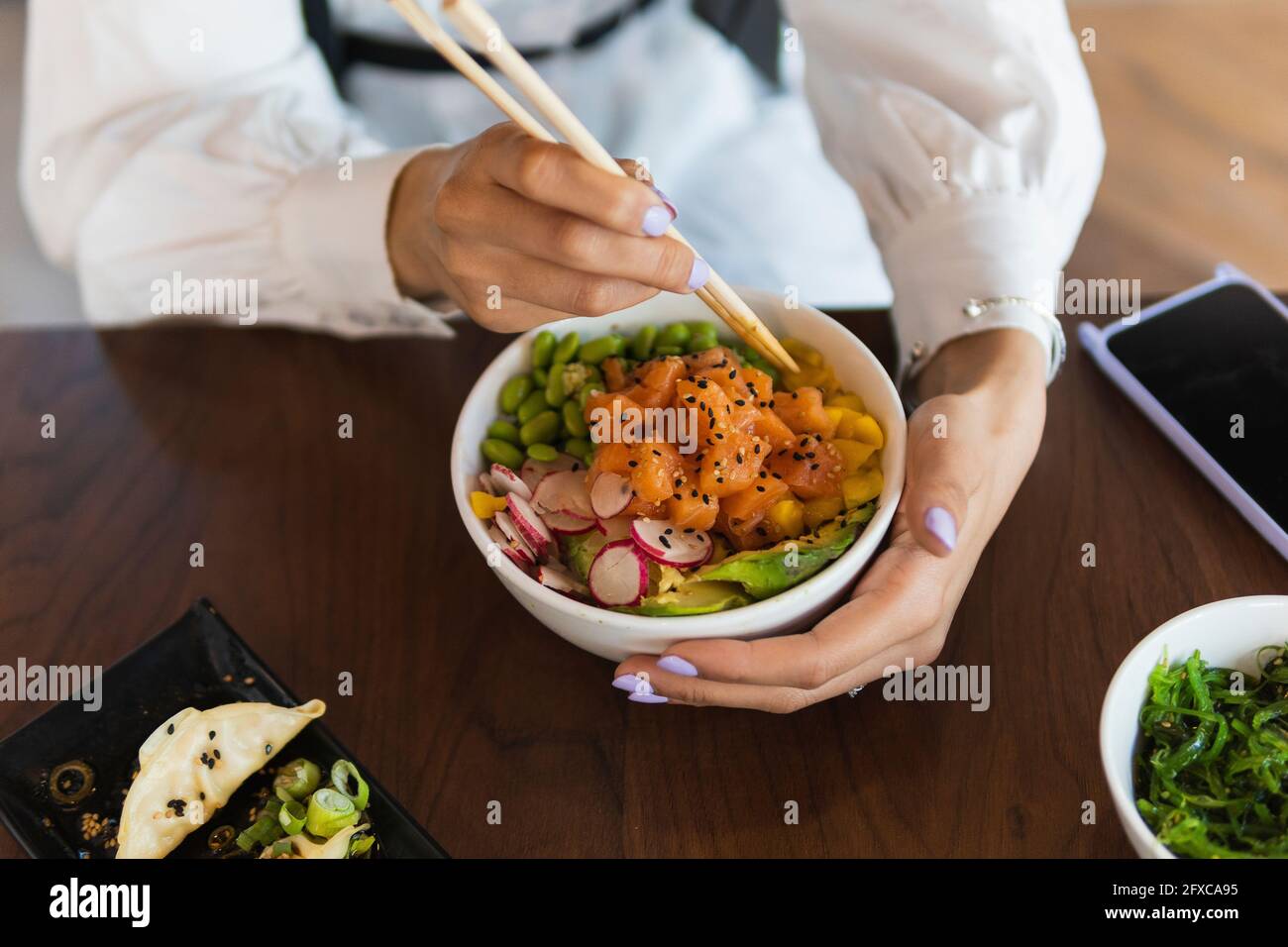Giovane donna che mangia una ciotola di poke con i bastoncini sul tavolo al ristorante Foto Stock