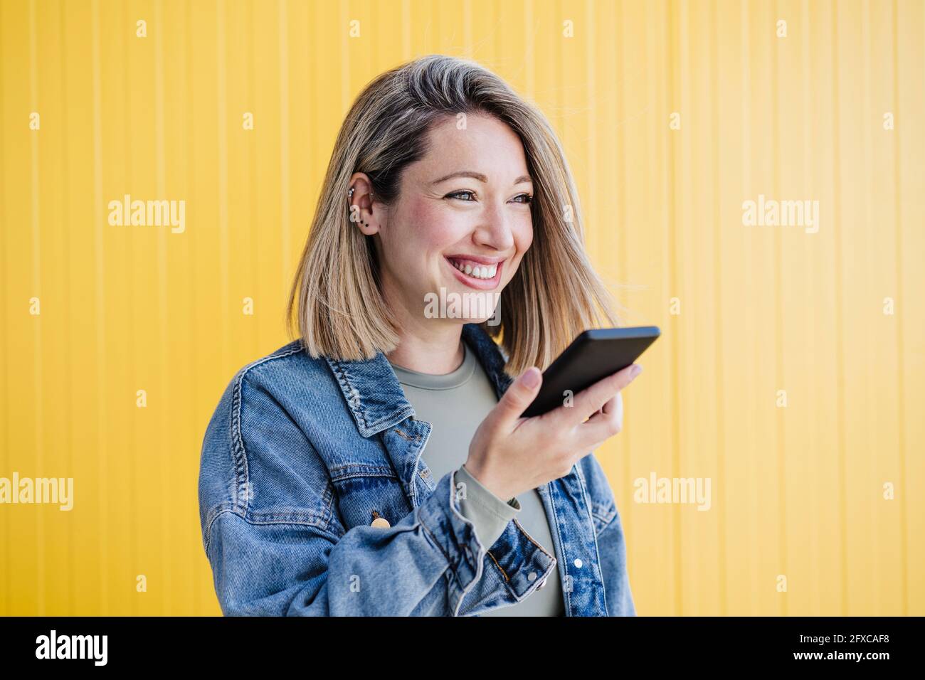 Donna felice che invia la segreteria telefonica tramite smartphone da parete gialla Foto Stock