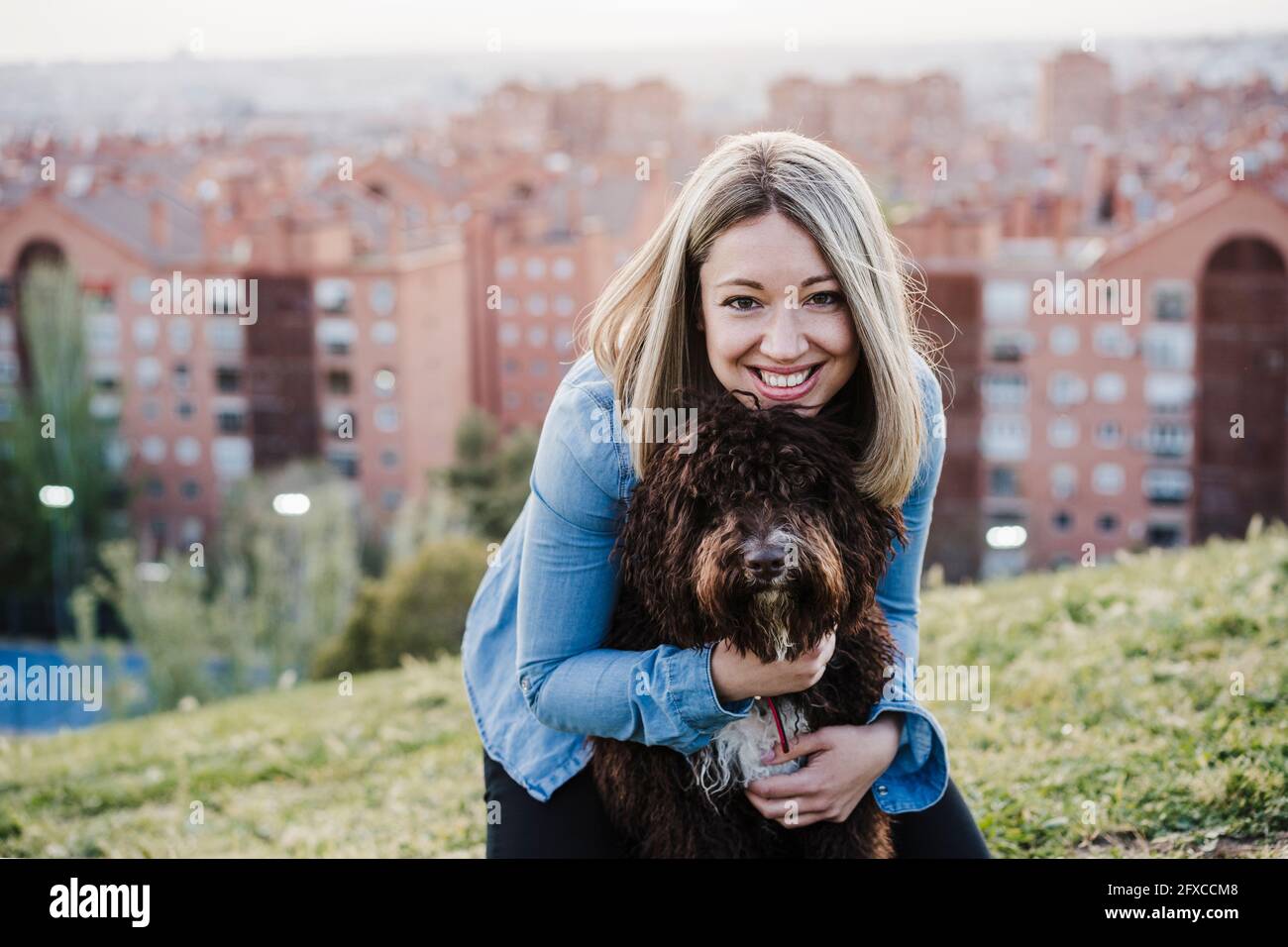 Donna sorridente con capelli biondi che abbracciano l'acqua spagnola cane su erba durante il tramonto Foto Stock
