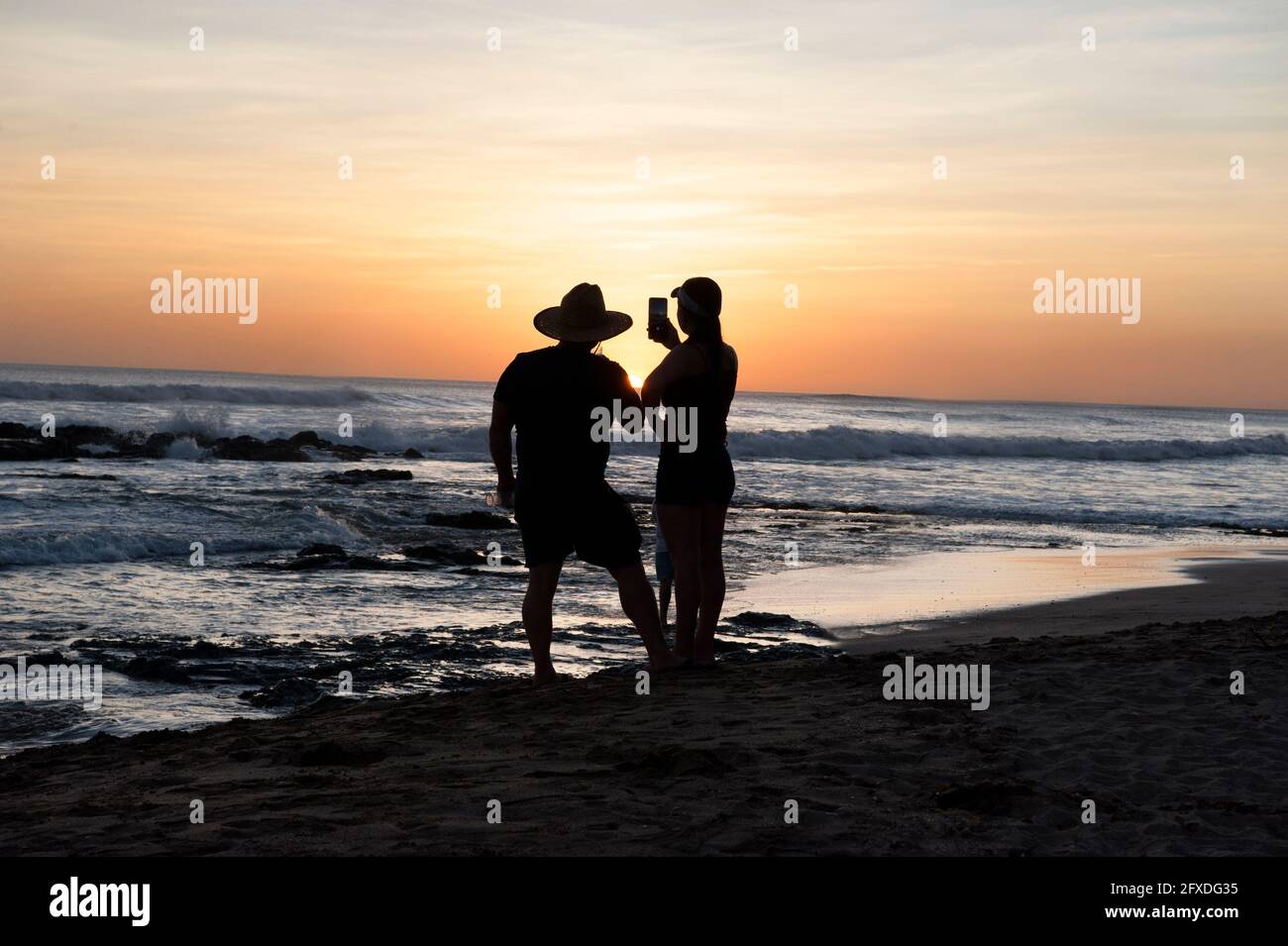 Una coppia scatta foto del tramonto con un telefono cellulare su una spiaggia in coppia guardando il tramonto da una spiaggia a Tamarindo, Costa Rica, America Centrale Foto Stock