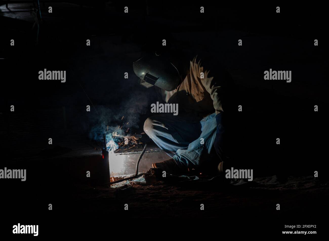 Un saldatore in jeans blu siede a gambe incrociate piegato sul suo lavoro nel buio della notte. Foto Stock