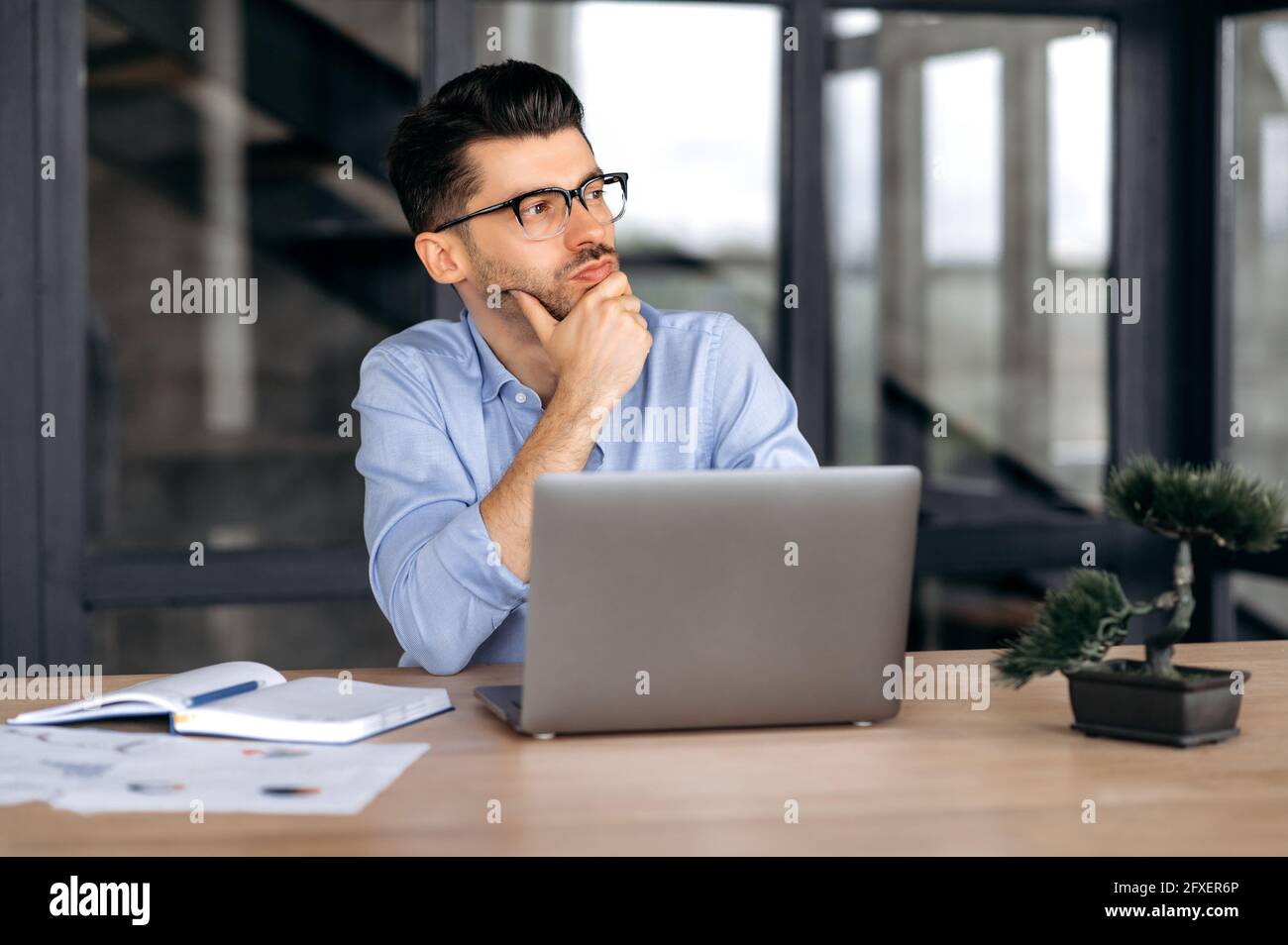 Pensivo successo bello giovane leader caucasico, manager o broker in abiti e bicchieri formali, seduto a un portatile in ufficio, guardando al lato, poggiando la testa sulla sua mano Foto Stock