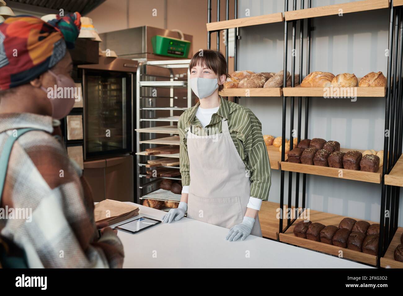 Giovane donna in uniforme e in maschera a cui si vende il pane il cliente nel panificio Foto Stock