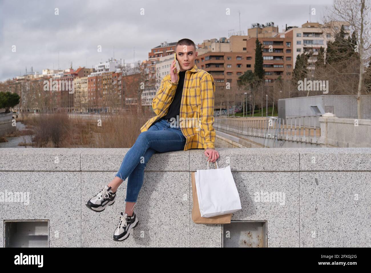 Giovane persona non binaria che parla sullo smartphone con le borse della spesa sedute su una parete. Make up persona androgina. Foto Stock