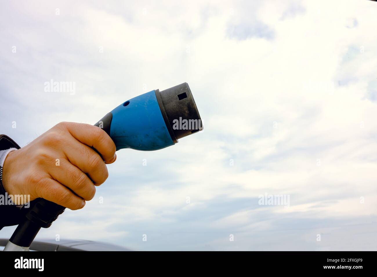 Uomo che tiene un caricabatterie EV contro il cielo nuvoloso. Copia spazio. Caricabatteria EV. Ricarica del veicolo elettrico. Energia alternativa. Tecnologia moderna. Qualità elevata Foto Stock