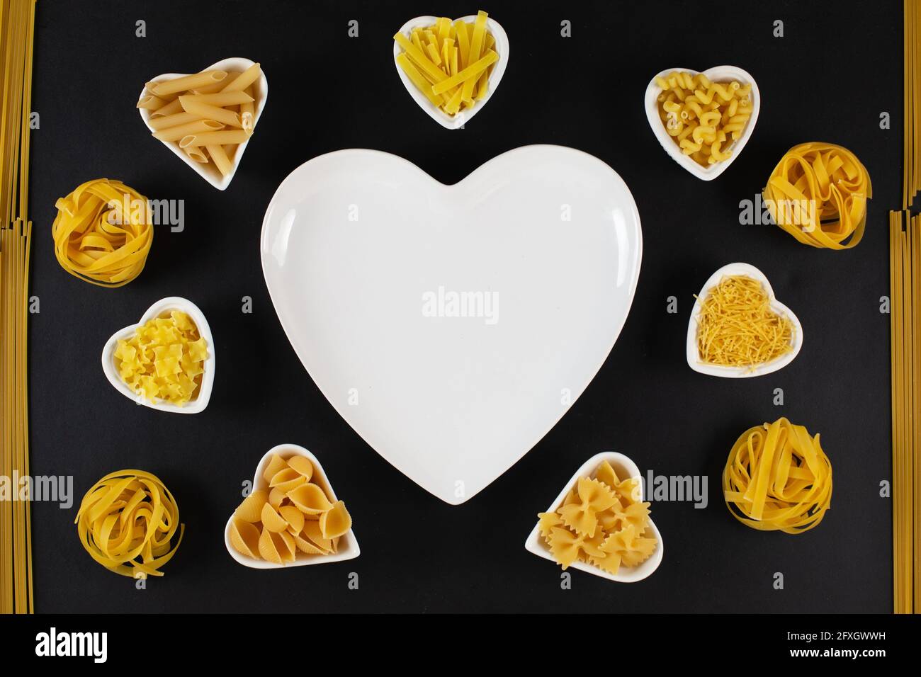 Selezione di pasta intorno a piatti di porcellana a forma di cuore su sfondo nero. Tagliatelle di pasta essiccate e non cotte. Concetto di pasta d'amore Foto Stock