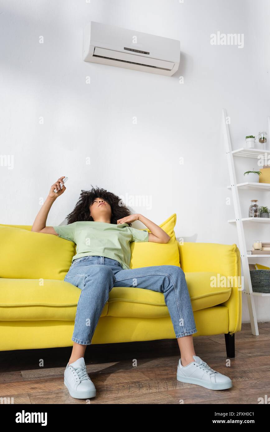 dispiaceva donna afroamericana che teneva il telecomando mentre era seduto  il divano giallo e sofferente di calore Foto stock - Alamy