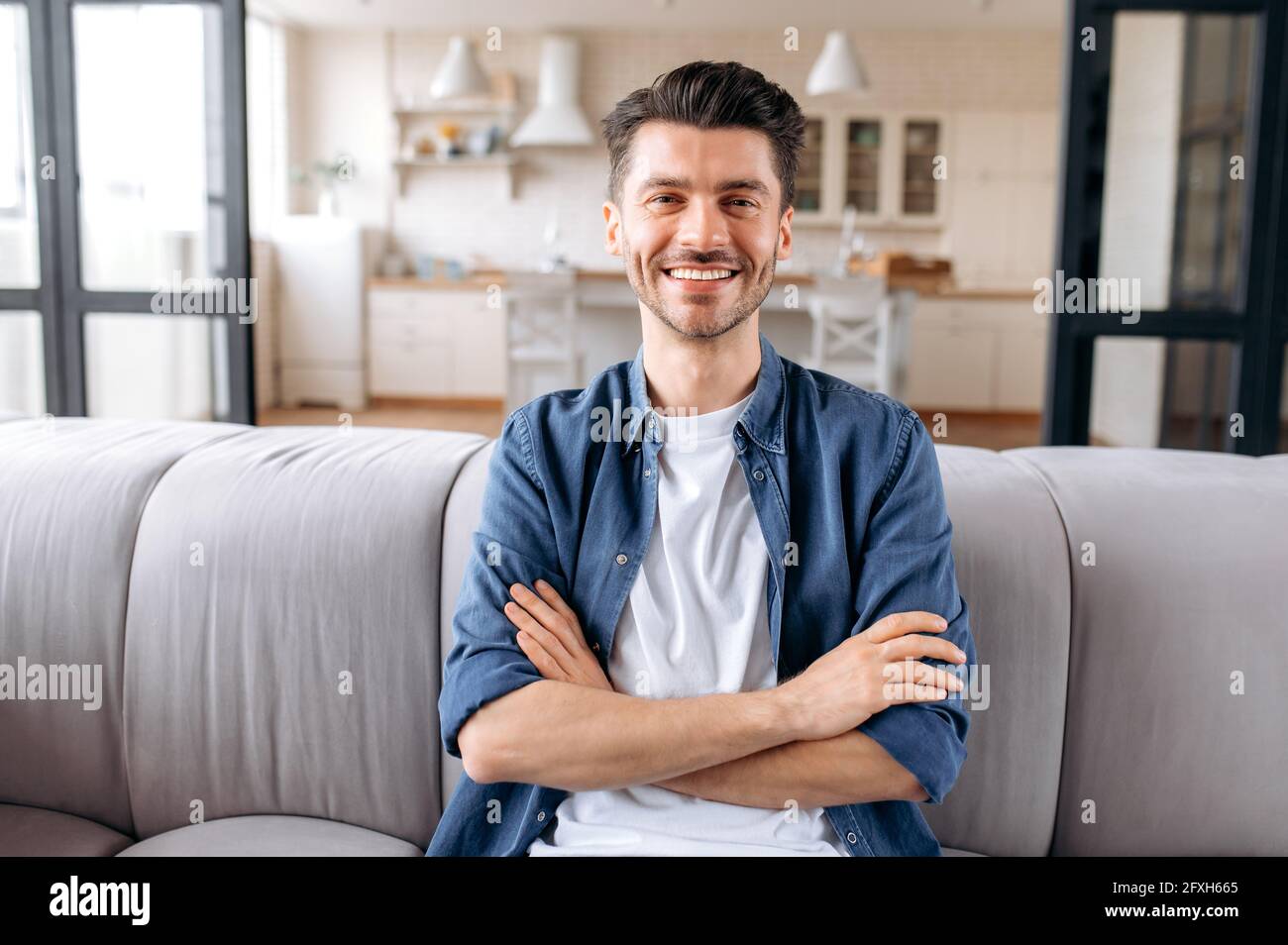 Ritratto di un attraente giovane caucasico, tranquillo e gioioso, seduto a casa con le braccia incrociate sul divano del soggiorno, indossando abiti casual di stile, guardando la macchina fotografica e sorridendo amichevole Foto Stock