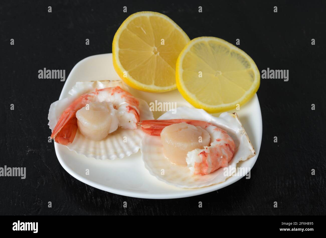 Code di gamberi bollite in una ciotola di ceramica con un capesante crudo nel guscio e limone su uno sfondo di legno scuro. Messa a fuoco selettiva. Foto Stock