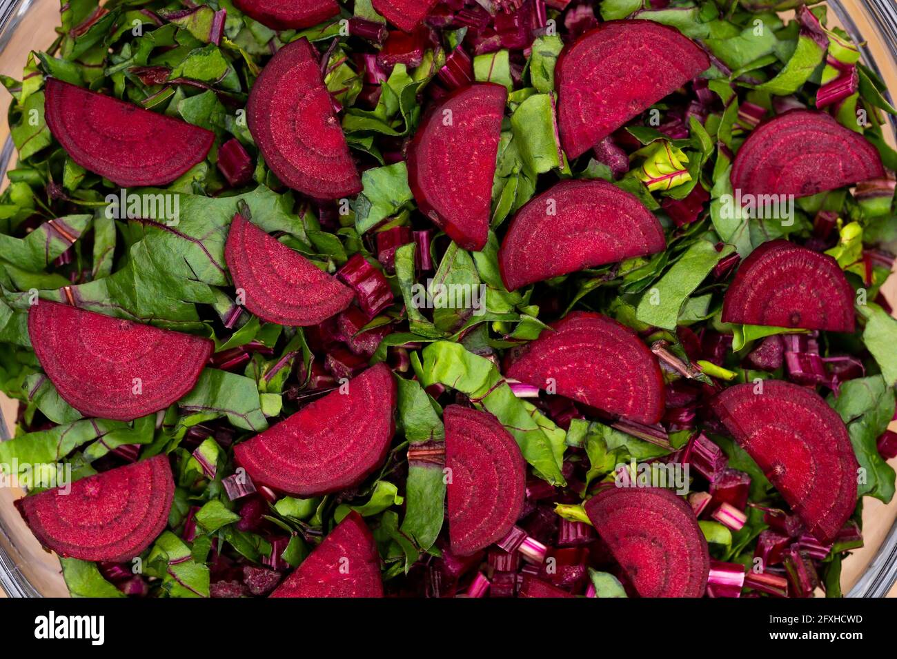 Foglie fresche tagliate e pezzi di barbabietole sistemati in una ciotola di vetro. Foto scattata in condizioni di luce artificiale e soffusa. Foto Stock