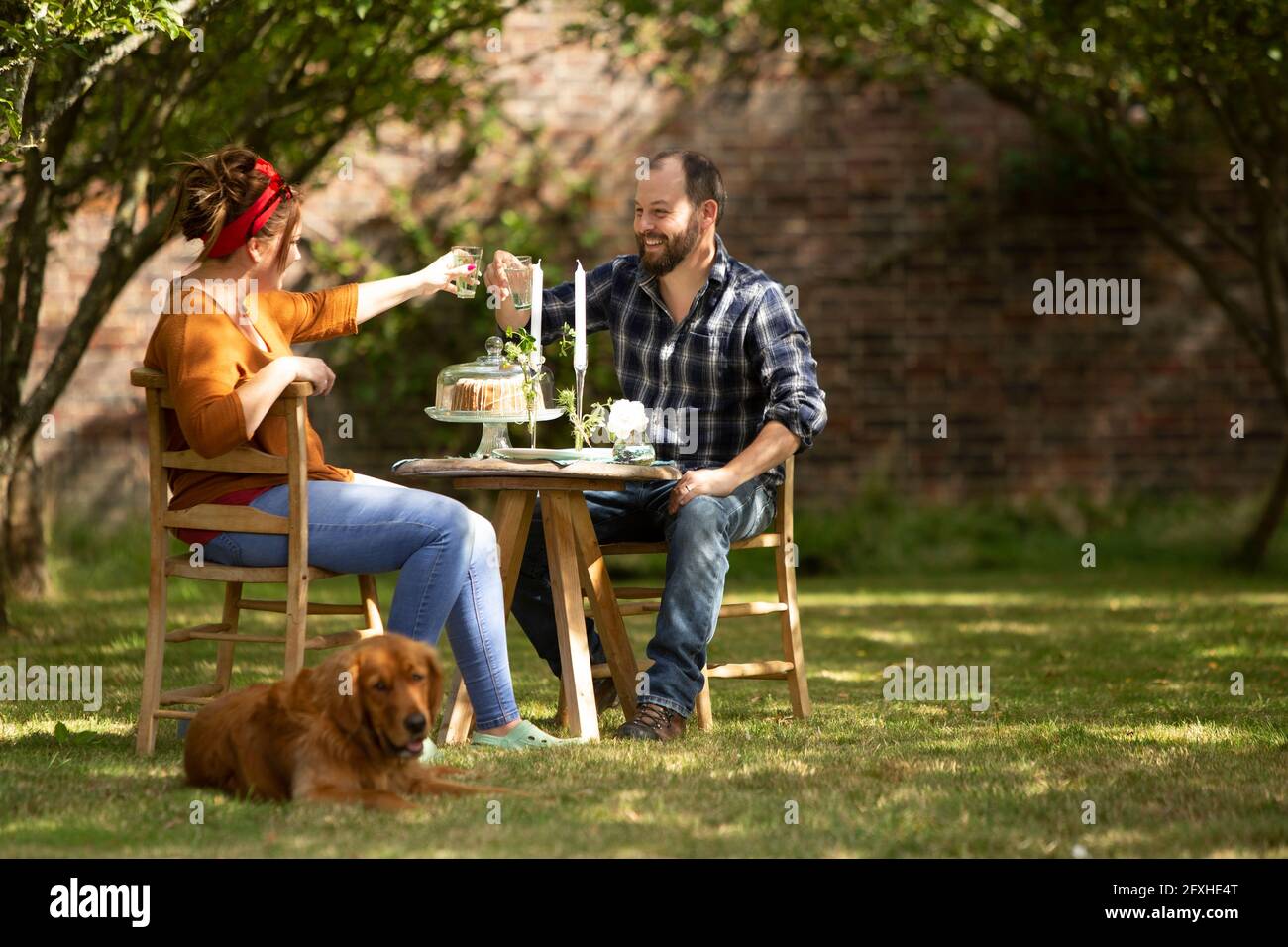 Felice coppia con i bicchieri da tostatura del cane al tavolo nel giardino estivo Foto Stock