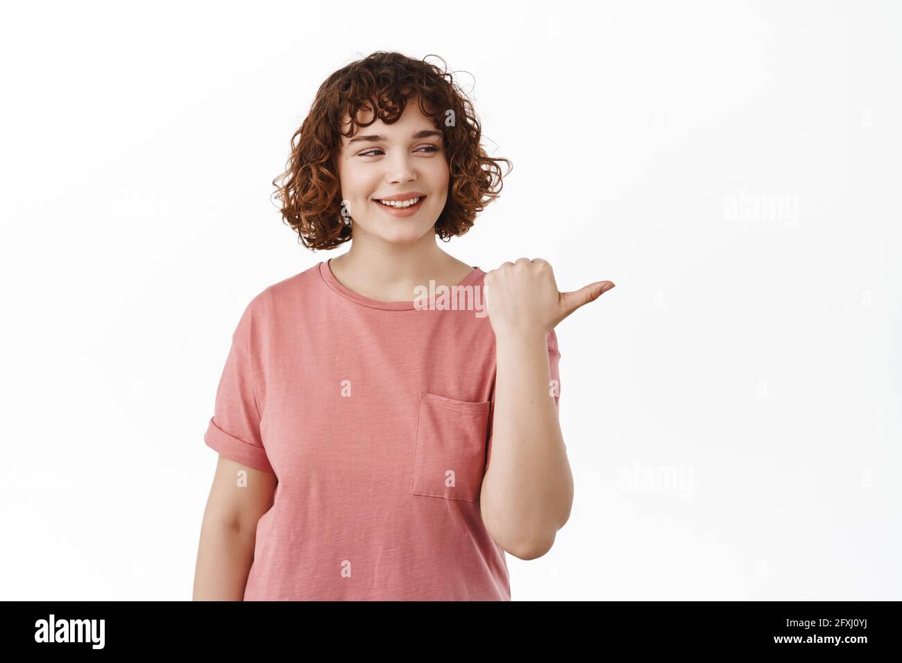 La giovane studentessa in t-shirt guarda a destra, punta il dito alla pubblicità con il volto speranzoso pensieroso, sorridente soddisfatto e felice, in piedi su bianco Foto Stock