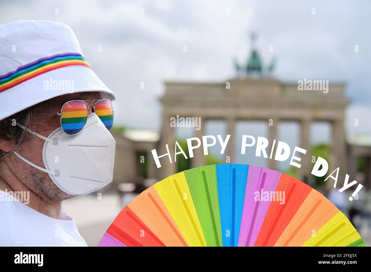 Uomo LGBT caucasico maturo a Berlino con ventola arcobaleno e nastro su cappello estivo bianco. Rainbow, simbolo dell'orgoglio gay LGBTQIA, la diversità. L'attivista maschile è Foto Stock