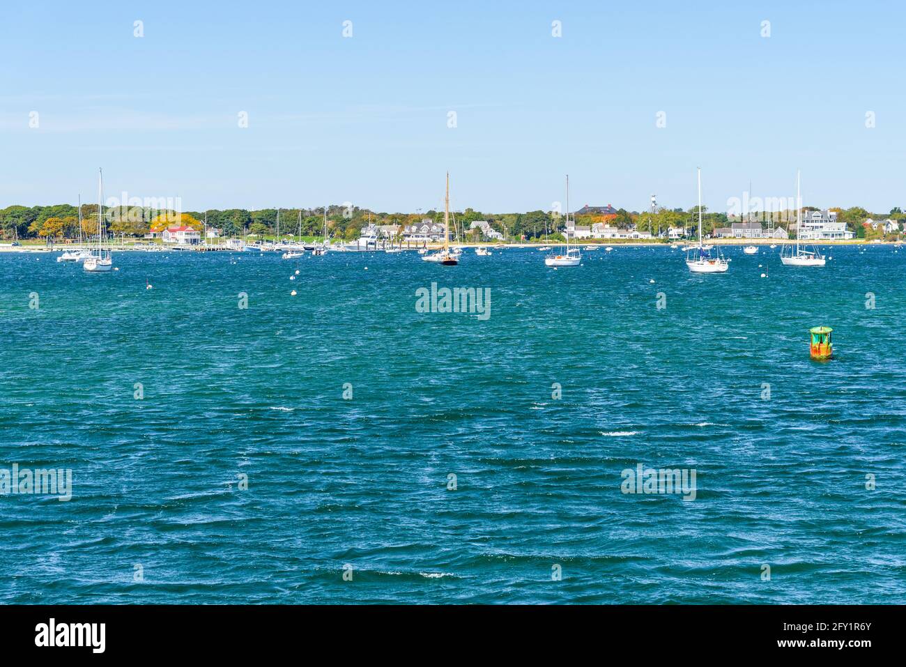 Barche a vela ancorate in acque blu al largo di una costa con case lungomare in un chiaro giorno d'autunno. Un indicatore verde canale buoy è in foregrond. Foto Stock