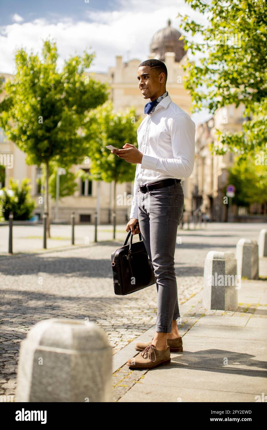Bel giovane uomo d'affari afro-americano che usa un telefono cellulare mentre attesa per un taxi su una strada Foto Stock