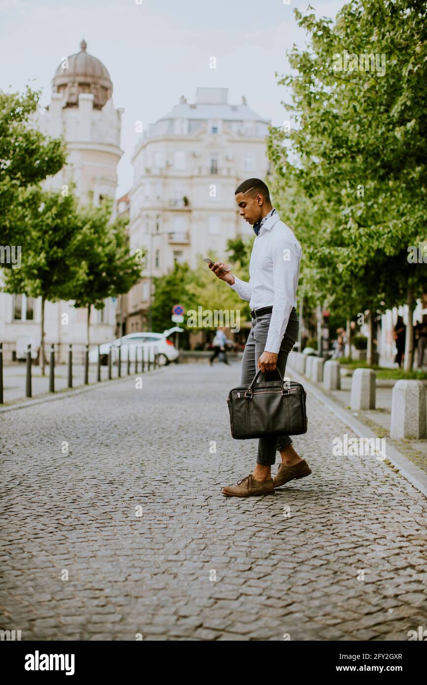 Bel giovane uomo d'affari afro-americano che usa un telefono cellulare mentre ceossing una strada Foto Stock