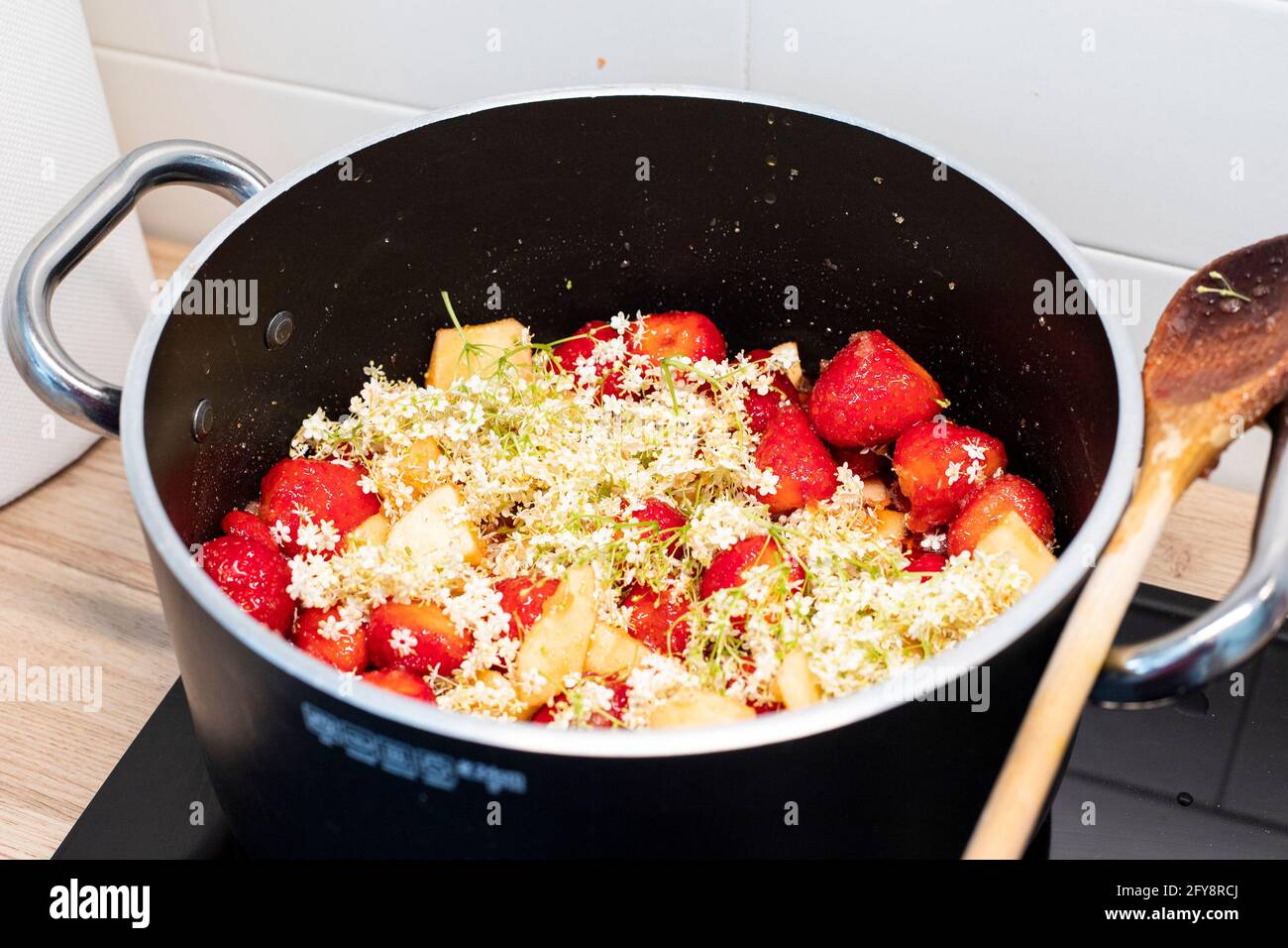 Fragole e fiori di sambuco marmellata cottura in pentola nera con cucchiaio di legno. La foraggazione primaverile conserva l'ispirazione della ricetta. Primo piano, vista laterale. Foto Stock