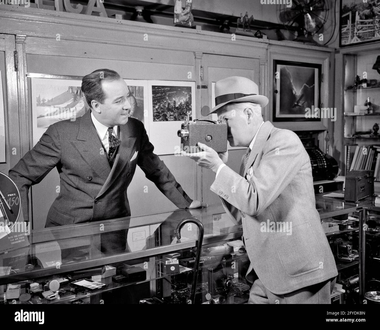 NEGOZIO DI FOTOCAMERE DEGLI ANNI '30 DEL 1940 CON UN CLIENTE UOMO CHE GUARDA ATTRAVERSO LA VISTA TROVA LA VIDEOCAMERA HOME MOVIE E IL VENDITORE DIETRO IL BANCONE - S10092 HAR001 HARS B&W UOMO DI MEZZA ETÀ ABILITÀ ACQUIRENTI ATTIVITÀ DIVERTENTE VENDITA HOBBY SERVIZIO CLIENTI INTERESSE E CONOSCENZA HOBBY PASSIME PIACERE OCCUPAZIONI NEGOZI HOME FILM FOTOGRAFIA COMMERCIO MEDIO-ADULTO MEDIO-ADULTO UOMO RELAX VENDITORI AMATEUR BIANCO E NERO IMPRESE ETNIA CAUCASICA GODIMENTO HAR001 VECCHIO STILE Foto Stock