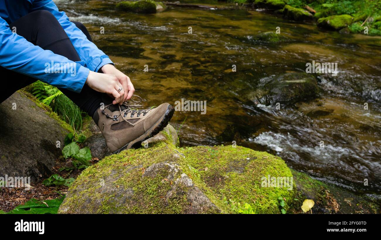 Donna seduta sulla roccia, fa allacciare lo stivaletto marrone che cammina seduto su una roccia coperta da muschio dal torrente di montagna piccolo fiume. Foto Stock