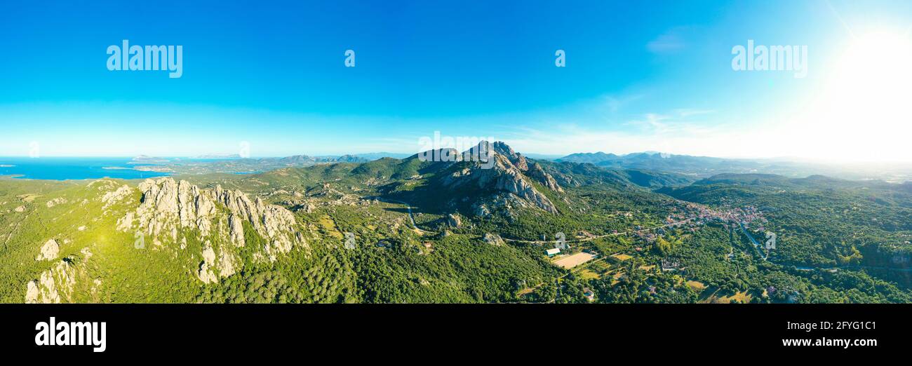 Vista dall'alto, splendida vista aerea del villaggio di San Pantaleo circondato da alcune montagne di granito, vegetazione verde e il Mar mediterraneo Foto Stock