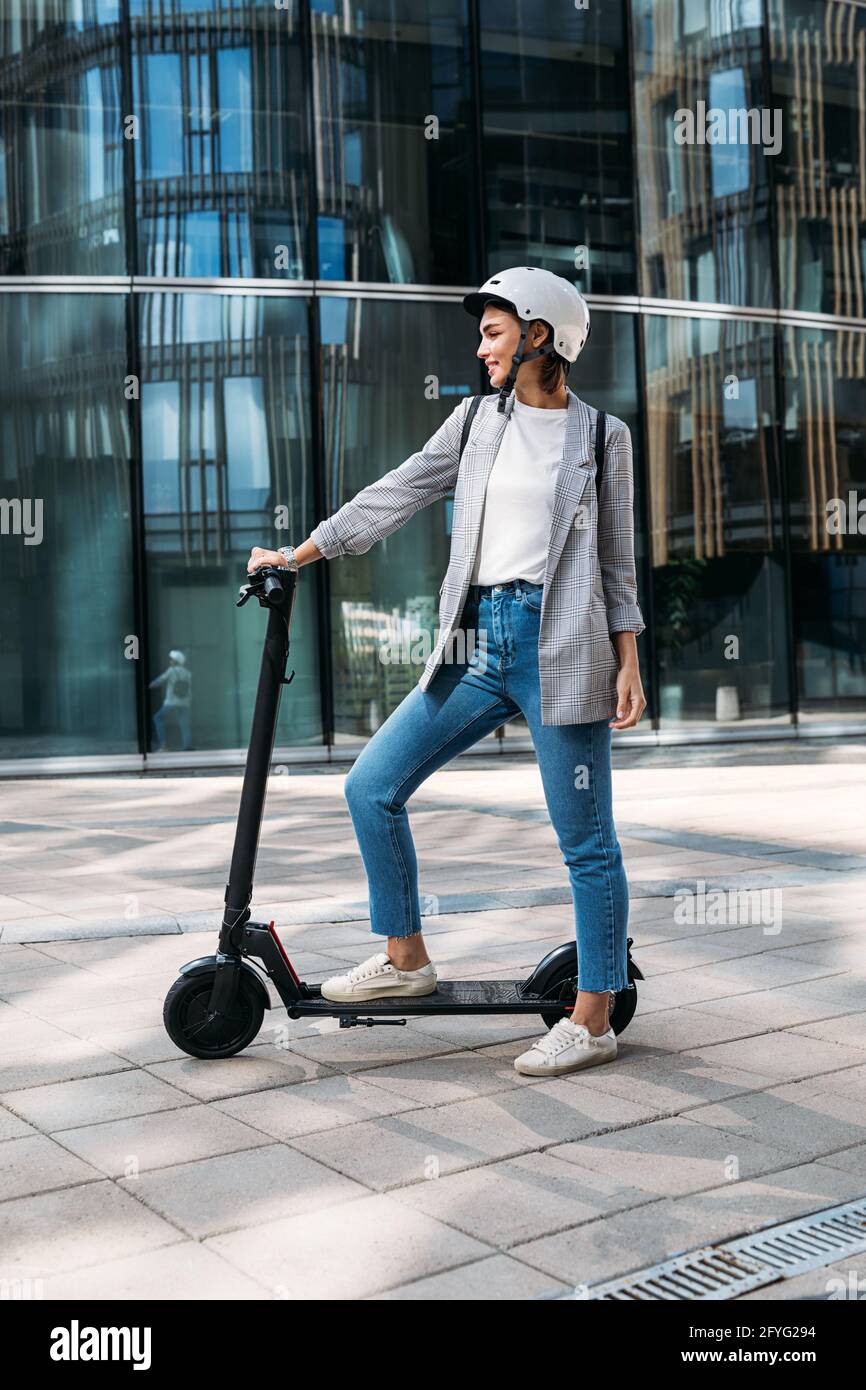 Vista laterale giovane donna in piedi su scooter elettrico vicino a un edificio di ufficio. Donna in casco pronta per un giro su scooter. Foto Stock
