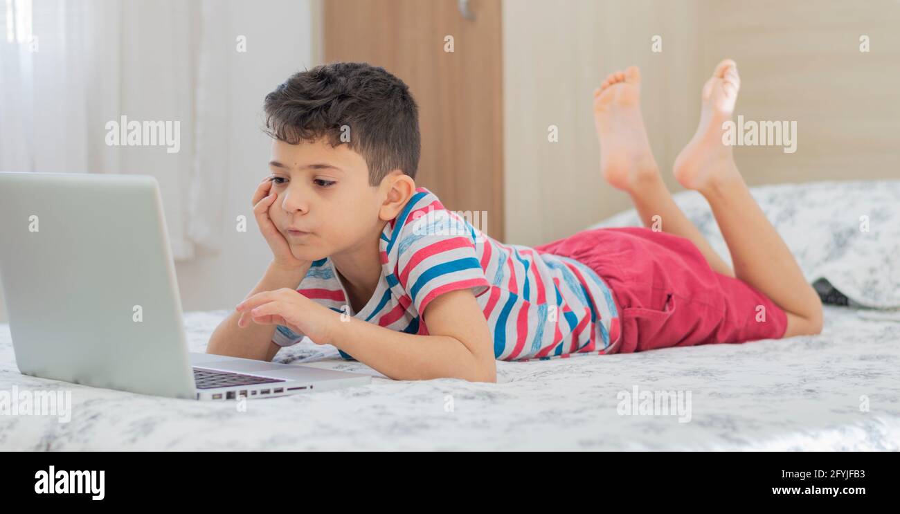 Bambino ragazzo sdraiato a letto usando un computer portatile. Lezione online sul video. Formazione remota a distanza. Nuovo studio scolastico normale. Bambini a scuola Foto Stock