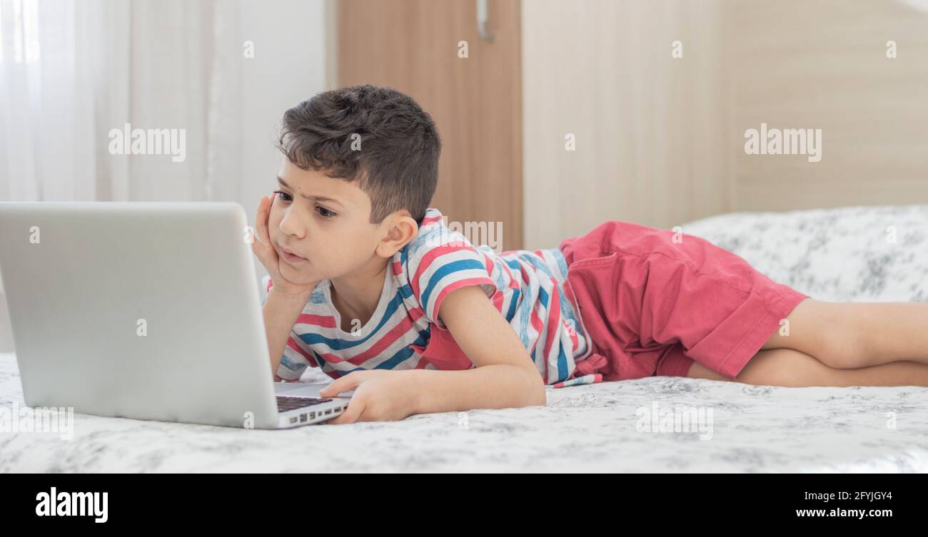 Bambino ragazzo sdraiato a letto usando un computer portatile. Lezione online sul video. Formazione remota a distanza. Nuovo studio scolastico normale. Bambini a scuola Foto Stock