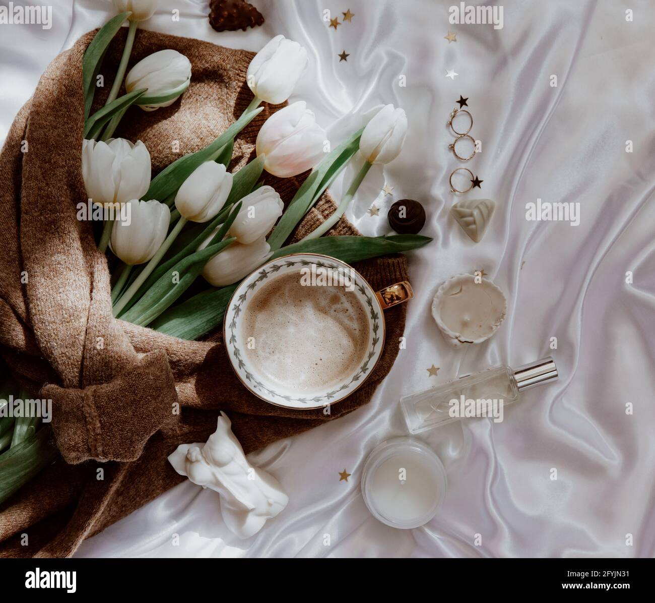 Mazzo di tulipani bianchi avvolti in un maglione di lana su una tovaglia di seta con candele, profumo e gioielli Foto Stock