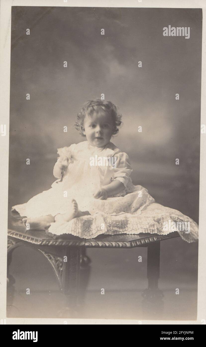 Cartolina fotografica d'epoca dei primi anni del XX secolo che mostra un bambino di 14 mesi seduto su un tavolo nella sua abito. Nominato Francis Wilfred Newman. Foto Stock