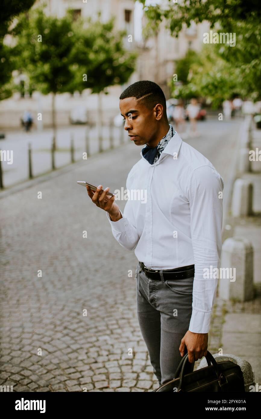 Bel giovane uomo d'affari afro-americano che usa un telefono cellulare mentre attesa di un taxi in attesa di un taxi su una strada Foto Stock