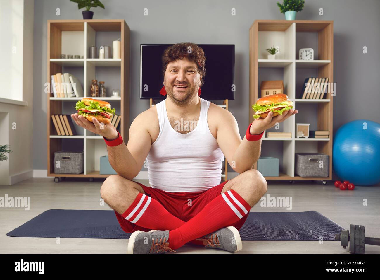 Felice uomo chubby seduto su tappeto sportivo, mangiare fast food e godere il suo giorno di cheat Foto Stock